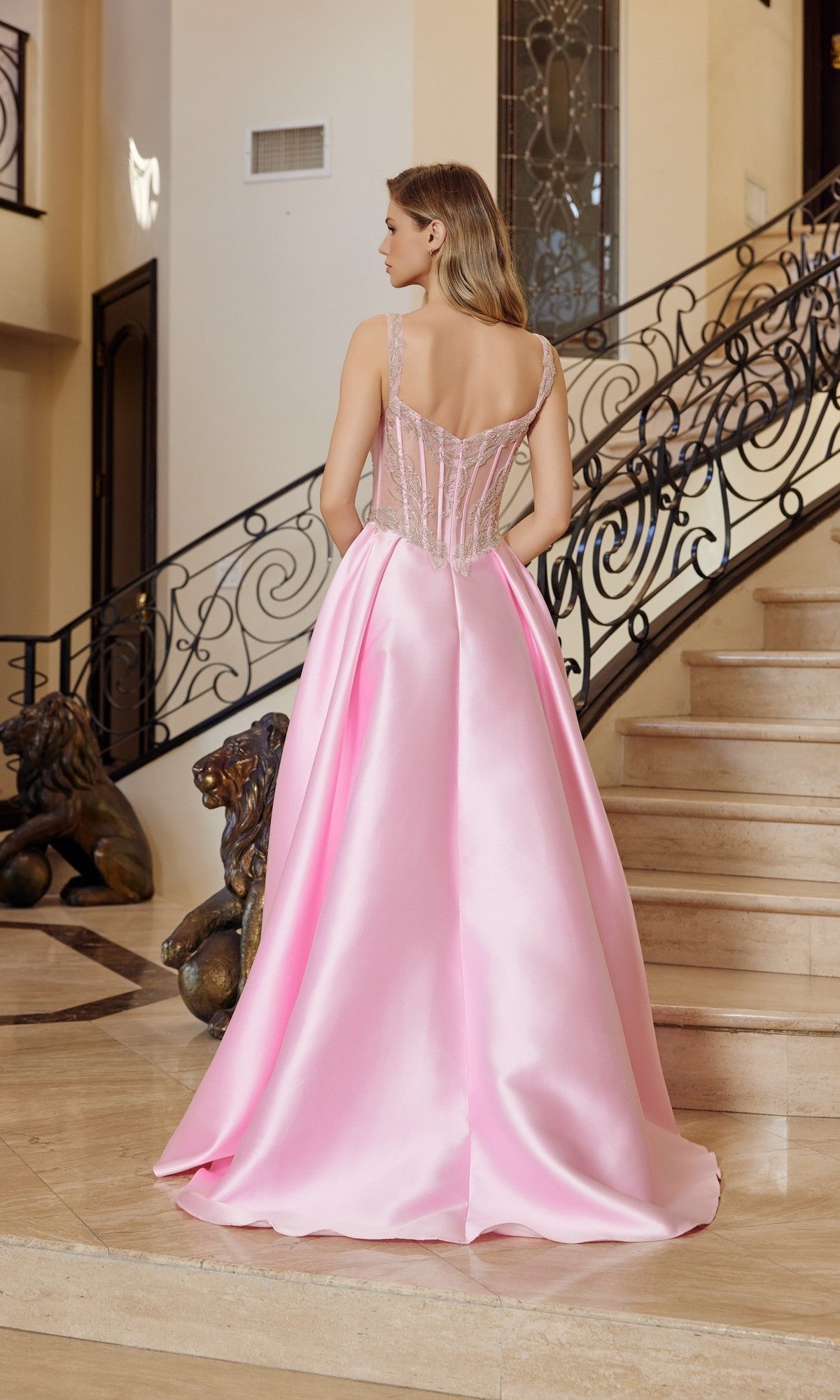 Wearing the Nox Anabel Q1775 Long Prom Dress in light pink satin, a woman with a fitted sheer corset bodice stands on marble stairs beside ornate railings and bronze lion statues.